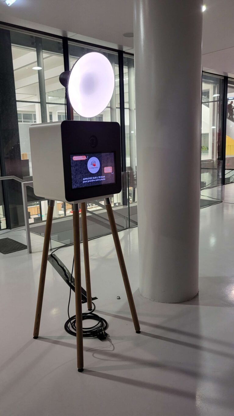 Borne selfie ONE, flash allumé, au pied d’une colonne dans un hall vitré