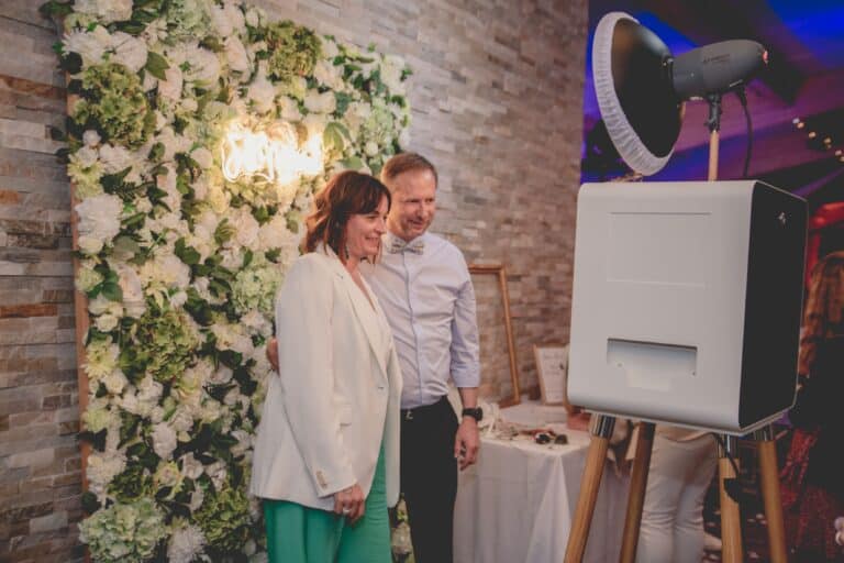 Un couple pose devant un mur de fleurs blanches, la borne selfie ONE au premier plan