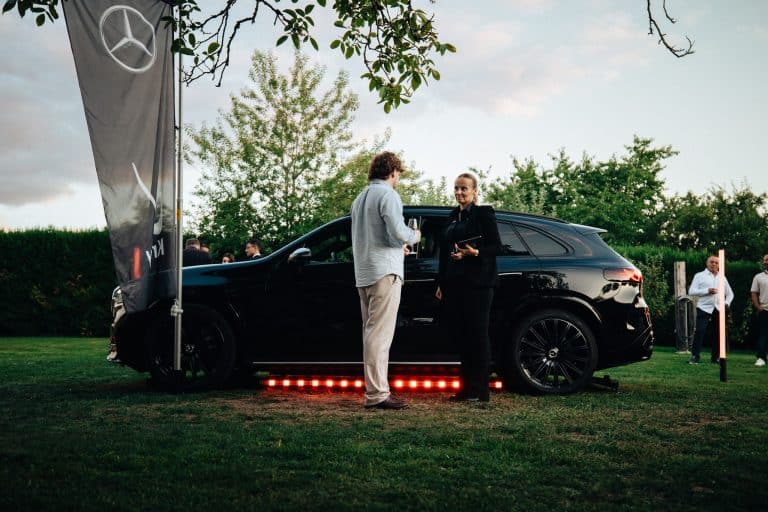 Deux personnes échangent devant un Mercedes GLC noir souligné de barres LED, drapeau Mercedes-Benz dans le jardin