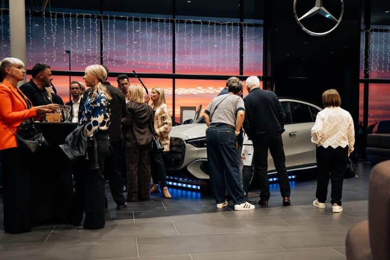 Invités en conversation devant la Mercedes Classe S, ciel rose du coucher de soleil derrière la baie vitrée du showroom