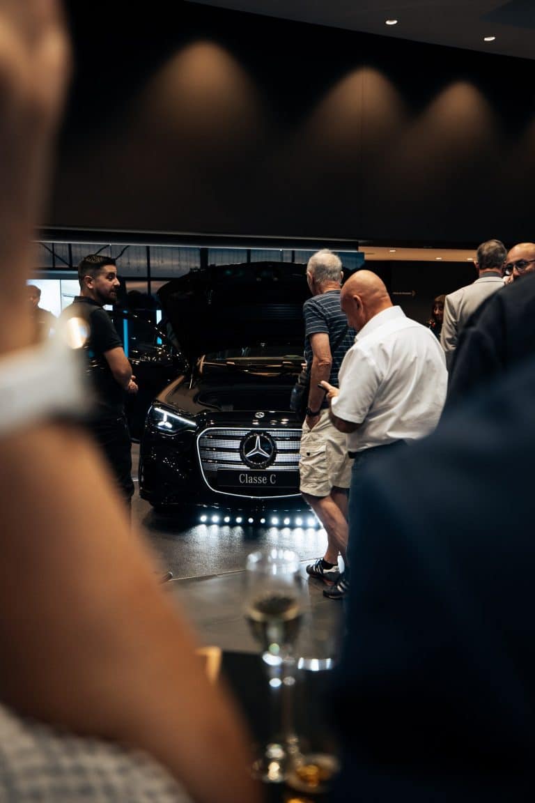 Calandre d'une Mercedes Classe C noire éclairée par une barre LED au sol, invités au premier plan, verre à la main