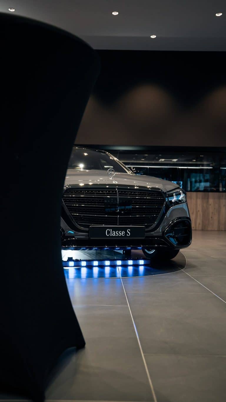 Mercedes Classe S vue de face derrière un mange-debout, barre LED bleue posée au sol sous la calandre, showroom sombre