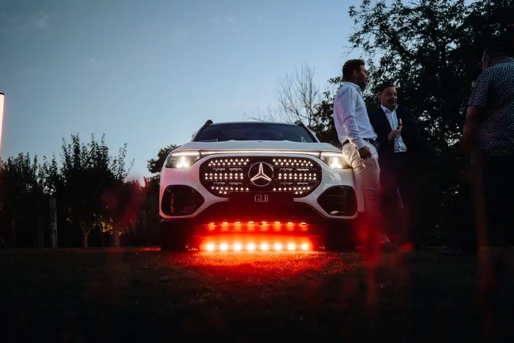 Face avant d'un Mercedes GLB blanc, calandre étoilée allumée et barre LED orangée posée sous le pare-chocs, invités à côté