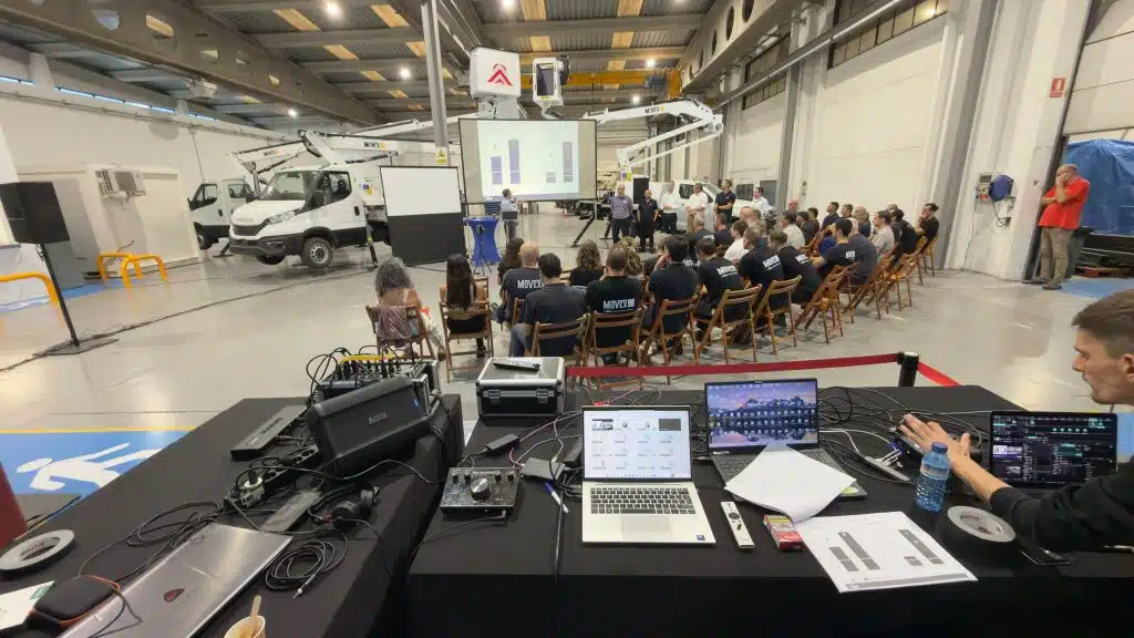 Régie vidéo LORENTS au premier plan, ordinateurs, console de mixage et enceinte, face aux équipes assises dans l'atelier devant les écrans de projection et une nacelle sur camion