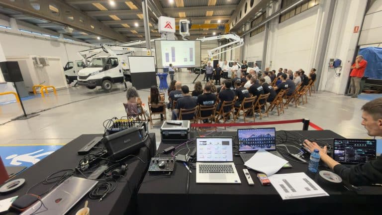 Régie vidéo LORENTS au premier plan, ordinateurs, console de mixage et enceinte, face aux équipes assises dans l'atelier devant les écrans de projection et une nacelle sur camion