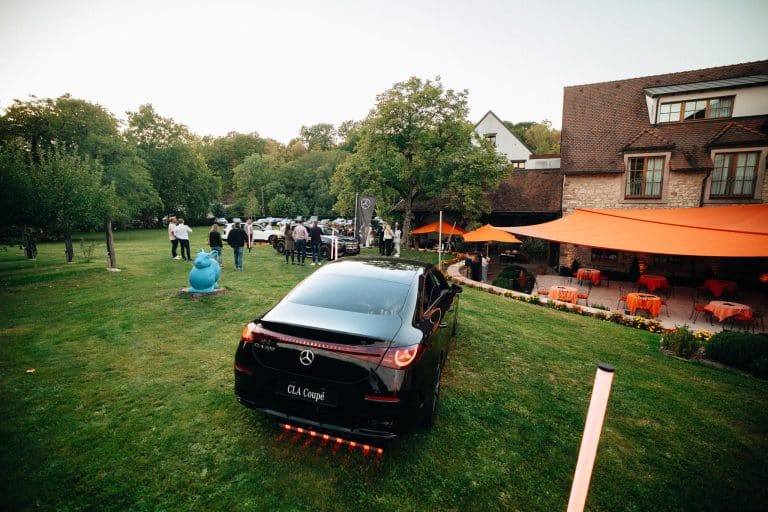 Jardin de l'Auberge Saint-Walfrid à Sarreguemines : Mercedes CLA Coupé sur la pelouse, terrasse aux parasols orange et invités