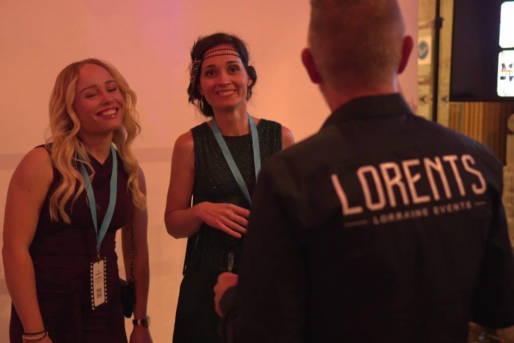 Le photographe LORENTS, de dos, fait poser deux invitées en soirée devant le fond du stand photo