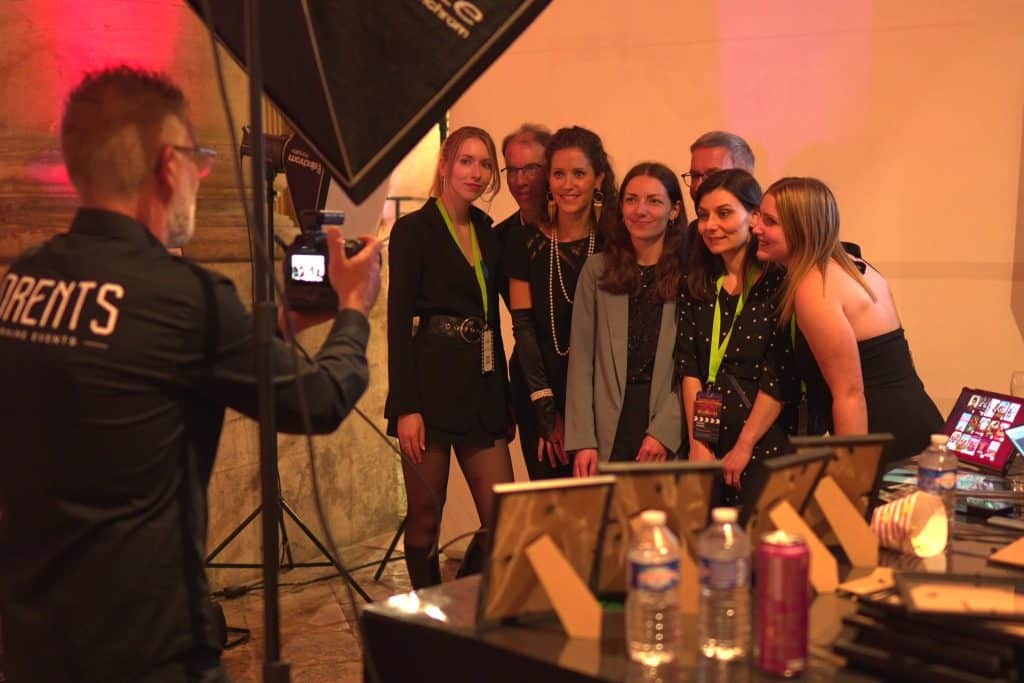 Le photographe LORENTS en veste siglée photographie un groupe d'invités devant le fond du stand photo, en soirée